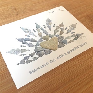 May include: A white card with a gray heart made of small stones in the center. The heart is surrounded by a sunburst design made of more small stones. The text "Start each day with a grateful heart" is printed on the card.