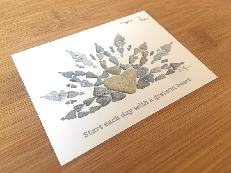 May include: A white card with a gray heart made of small stones in the center. The heart is surrounded by a sunburst design made of more small stones. The text "Start each day with a grateful heart" is printed on the card.