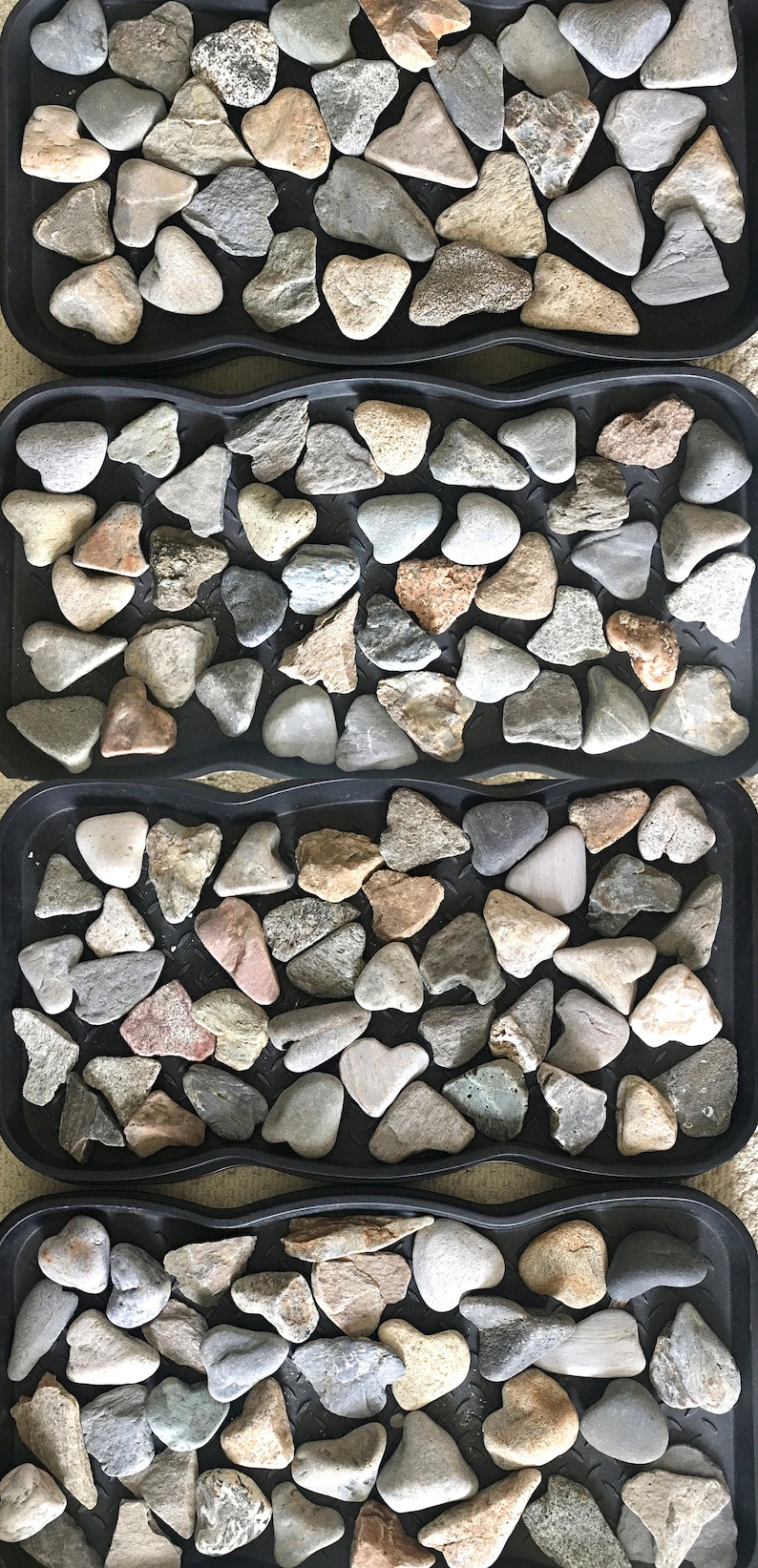 May include: Four black trays filled with smooth, gray, brown, and white heart-shaped stones. The stones are arranged in a pattern.