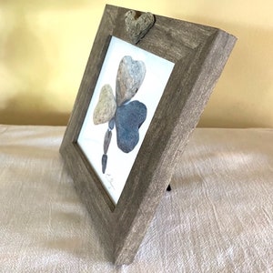 May include: A framed artwork featuring a clover made of stones. The clover is made of four stones, three of which are heart-shaped. The frame is a light brown wood with a distressed finish. The artwork is signed by the artist.