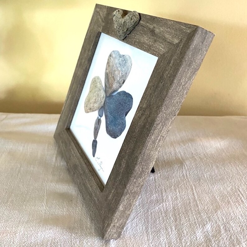 May include: A framed artwork featuring a clover made of stones. The clover is made of four stones, three of which are heart-shaped. The frame is a light brown wood with a distressed finish. The artwork is signed by the artist.