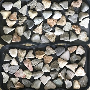 May include: Four black trays filled with smooth, gray, brown, and white heart-shaped stones. The stones are arranged in a pattern.