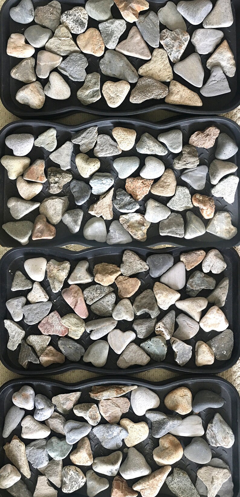 May include: Four black trays filled with smooth, gray, brown, and white heart-shaped stones. The stones are arranged in a pattern.