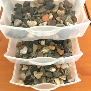 May include: Three clear plastic drawers filled with a variety of smooth, gray, brown, and tan stones. The drawers are stacked on top of each other.