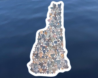 May include: A state outline of New Hampshire created with small, heart-shaped stones in shades of gray, brown, and white. The outline is set against a blue background.