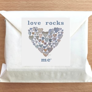 May include: A white envelope with a card inside. The card has a heart shape made of small gray rocks with the text "love rocks me" in blue.