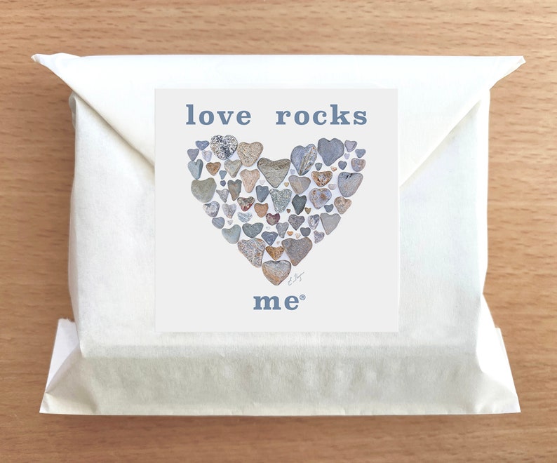 May include: A white envelope with a card inside. The card has a heart shape made of small gray rocks with the text "love rocks me" in blue.