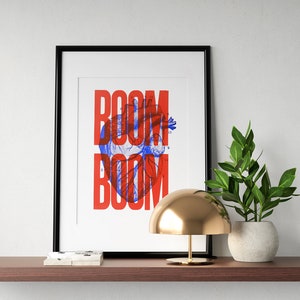 Może przedstawiać: Czarny obraz w ramie z napisem "BOOM BOOM" w czerwonych i niebieskich literach. Litery są nałożone na vintage'ową ilustrację anatomiczną ludzkiego serca. Obraz jest umieszczony na półce z złotą lampą i doniczką z rośliną.
