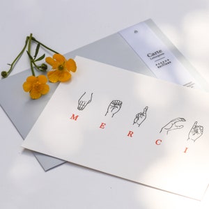 May include: A white card with the word "MERCI" spelled out in sign language. The letters are in red and the hands are in black. The card is on top of a light gray envelope with the words "Carte Letterpress Pappus Editions" printed on it.