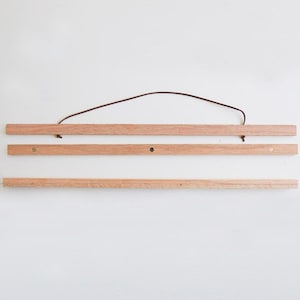 K&ouml;nnte beinhalten: Ein Holz-Posterhalter mit einem braunen Lederband. Der Halter besteht aus drei Holzst&uuml;cken und messingfarbenen Metallhaken an jedem Ende.