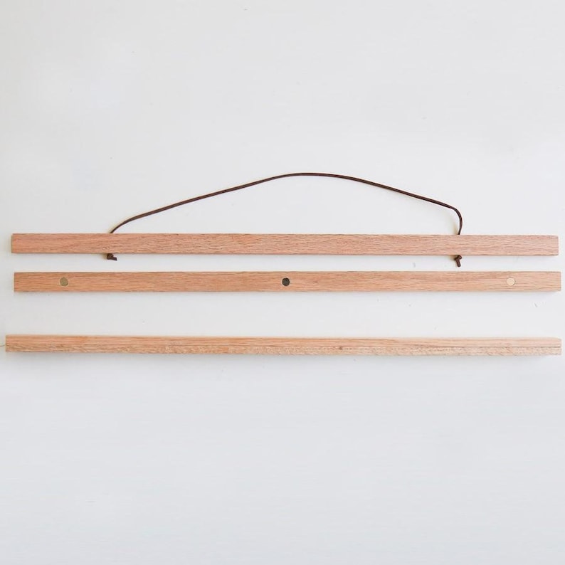 K&ouml;nnte beinhalten: Ein Holz-Posterhalter mit einem braunen Lederband. Der Halter besteht aus drei Holzst&uuml;cken und messingfarbenen Metallhaken an jedem Ende.