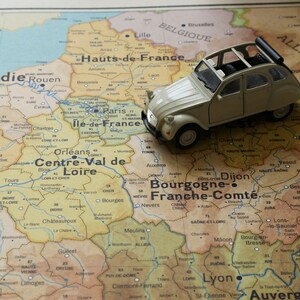 May include: A toy car, a light beige Citro&euml;n 2CV, is placed on a vintage map of France. The car is positioned over the region of Burgundy, Franche-Comt&eacute;, with the city of Dijon highlighted.