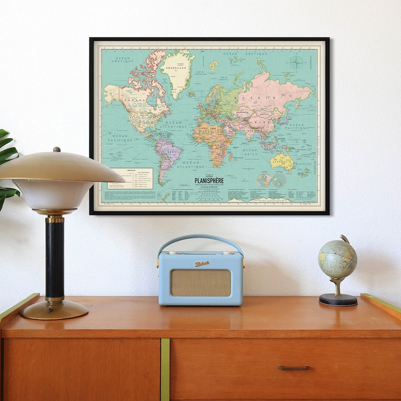 Op de afbeelding: Een vintage wereldkaartprint in een zwart frame hangt aan een witte muur boven een houten dressoir. Een blauwe radio en een kleine globe staan op het dressoir.