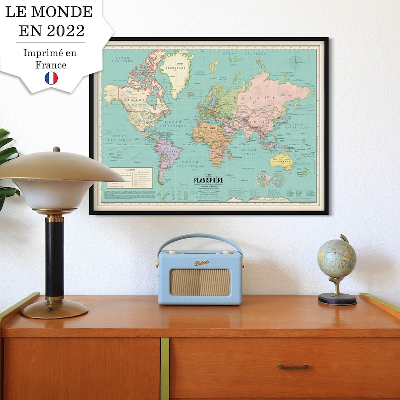 Op de afbeelding: Een vintage wereldkaartprint in een zwart frame, hangend aan een witte muur boven een houten dressoir. Een blauwe radio en een kleine globe staan op het dressoir.