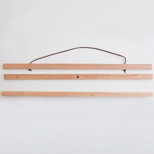 May include: A wooden poster hanger with a brown leather cord. The hanger has three pieces of wood and brass-coloured metal hooks on each end.