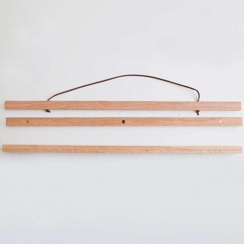 May include: A wooden poster hanger with a brown leather cord. The hanger has three pieces of wood and brass-coloured metal hooks on each end.
