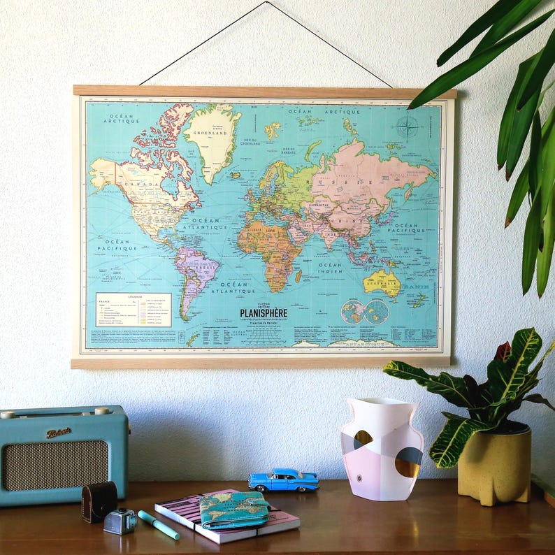 K&ouml;nnte beinhalten: Ein Vintage-Weltkarten-Druck mit blauem Hintergrund und bunten Kontinenten. Die Karte ist mit einem Holzrahmen gerahmt und h&auml;ngt an einer hellblauen Wand. Auf einem Holztisch stehen eine Pflanze in einem gelben Topf, ein blaues Spielzeugauto, eine rosa-goldene Vase und ein blaues Radio.