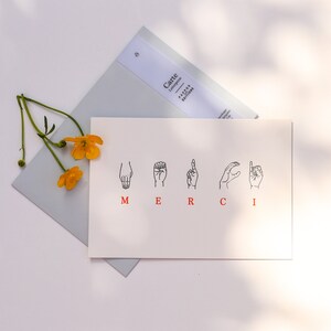 May include: A white card with the word "MERCI" spelled out in red letters. Each letter is represented by a hand in American Sign Language. The card is on top of a light blue envelope with the text "Carte Les Impressions" on it.