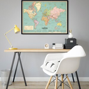 Op de afbeelding: Een vintage-stijl wereldkaart in een zwart frame hangt aan een lichtgrijze muur boven een houten bureau met een witte stoel. De kaart is op een lichtblauwe achtergrond met landen die in verschillende kleuren zijn omlijnd. De kaart heeft de titel "Planisphere".