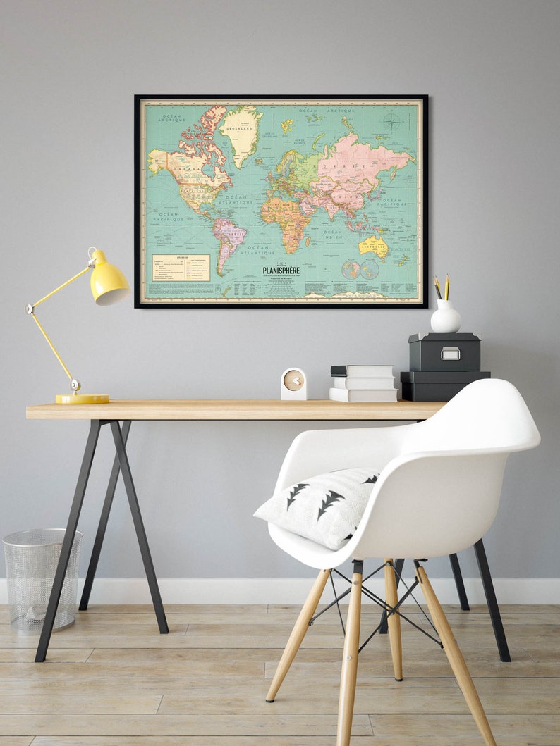 Op de afbeelding: Een vintage-stijl wereldkaart in een zwart frame hangt aan een lichtgrijze muur boven een houten bureau met een witte stoel. De kaart is op een lichtblauwe achtergrond met landen die in verschillende kleuren zijn omlijnd. De kaart heeft de titel "Planisphere".