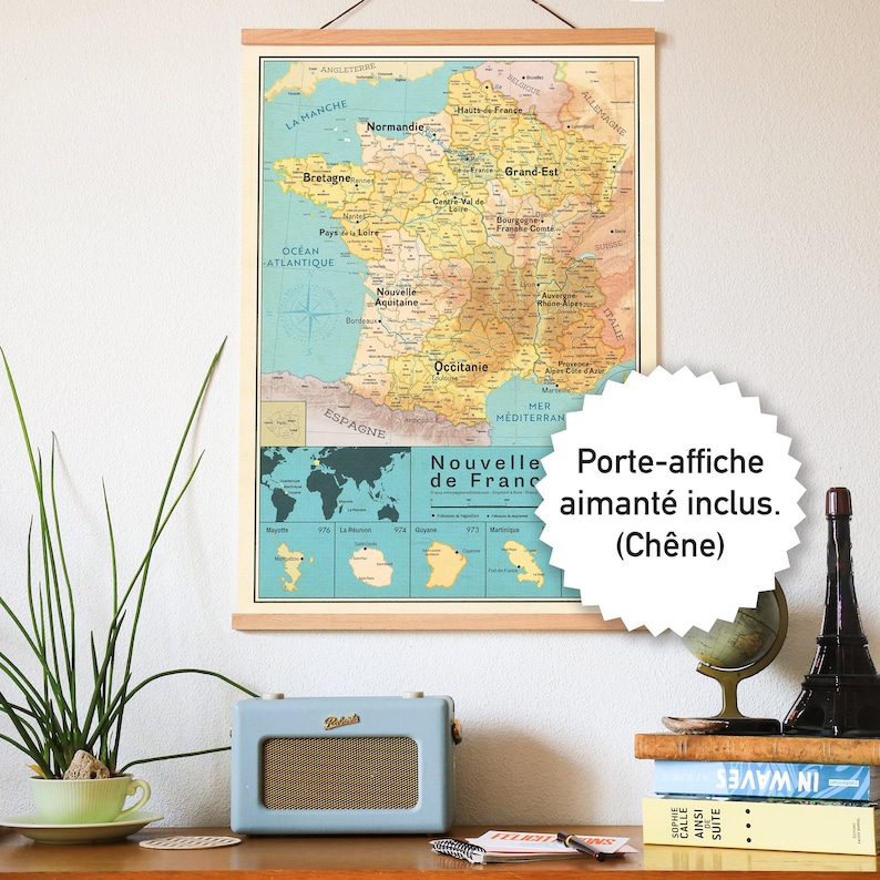 May include: A vintage-style map of France with a wooden frame and a hanging system. The map is printed on a light blue background with brown and yellow accents. The map includes the regions of France and a smaller map of the world showing the overseas territories. The text "Nouvelle de France" is printed in a brown font on the map. The map is displayed on a table with a radio, a cup, a plant, and other decorative items.