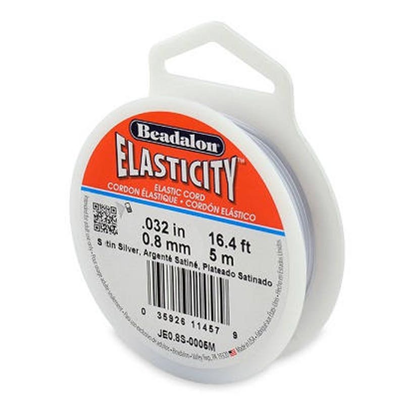 May include: A spool of silver-colored Beadalon Elasticity elastic cord. The label reads "Beadalon Elasticity Elastic Cord, 0.032 in, 0.8 mm, 16.4 ft, 5 m, Satin Silver, Argent&eacute; Satin&eacute;, Plateado Satinado, 35926 11457, JE0.8S-0006M".