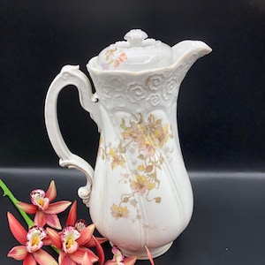 May include: White porcelain coffee pot with a floral design and a lid. The pot has a handle and a spout. The pot is sitting on a black surface with red orchids.