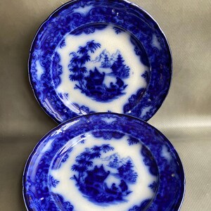 May include: Two blue and white porcelain plates with a blue and white Asian-inspired design. The plates have a scalloped edge and a white center with a blue design of a pagoda, trees, and figures in a boat.