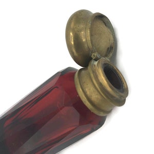 May include: A red glass bottle with a brass lid and a cotton swab inside. The bottle is faceted and has a vintage look.
