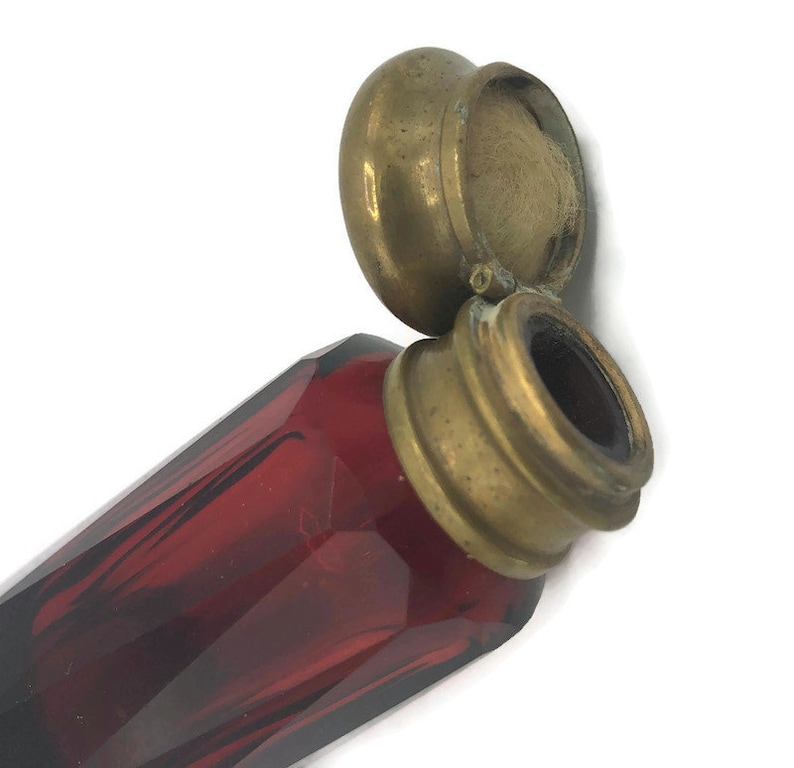 May include: A red glass bottle with a brass lid and a cotton swab inside. The bottle is faceted and has a vintage look.
