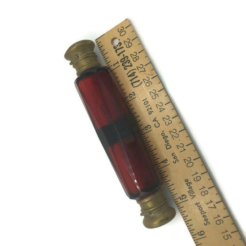 May include: A red glass bottle with gold metal caps. The bottle is approximately 3 inches long and has a faceted design. A wooden ruler is in the background.
