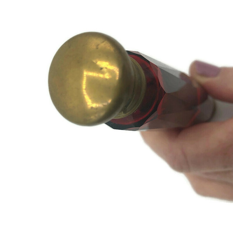 May include: A small, red glass bottle with a gold metal cap. The bottle is faceted and has a rounded shape. The cap is round and has a smooth surface.