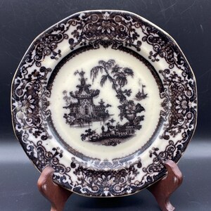 May include: A black and white porcelain plate with a decorative border and a scene of a pagoda and trees in the center.