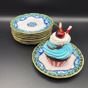 May include: A blue and white cupcake with chocolate frosting and a red cherry on top, sitting on a white plate with a blue and green floral design. The cupcake is surrounded by a stack of blue and white plates with gold trim.