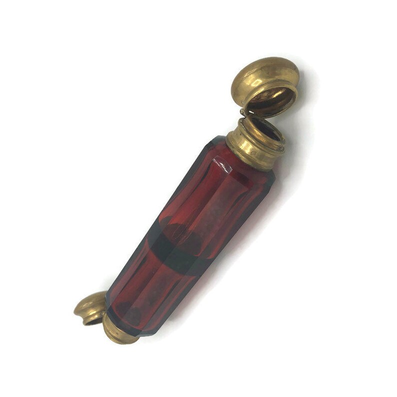 May include: A small, red glass bottle with a brass lid and a brass cap on the bottom. The bottle has a faceted design.