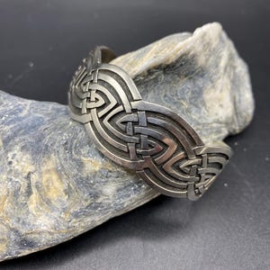 May include: A silver-toned cuff bracelet with an intricate Celtic knot design. The bracelet is curved and features interwoven patterns, creating a textured surface. The bracelet is displayed on a gray rock against a dark background.