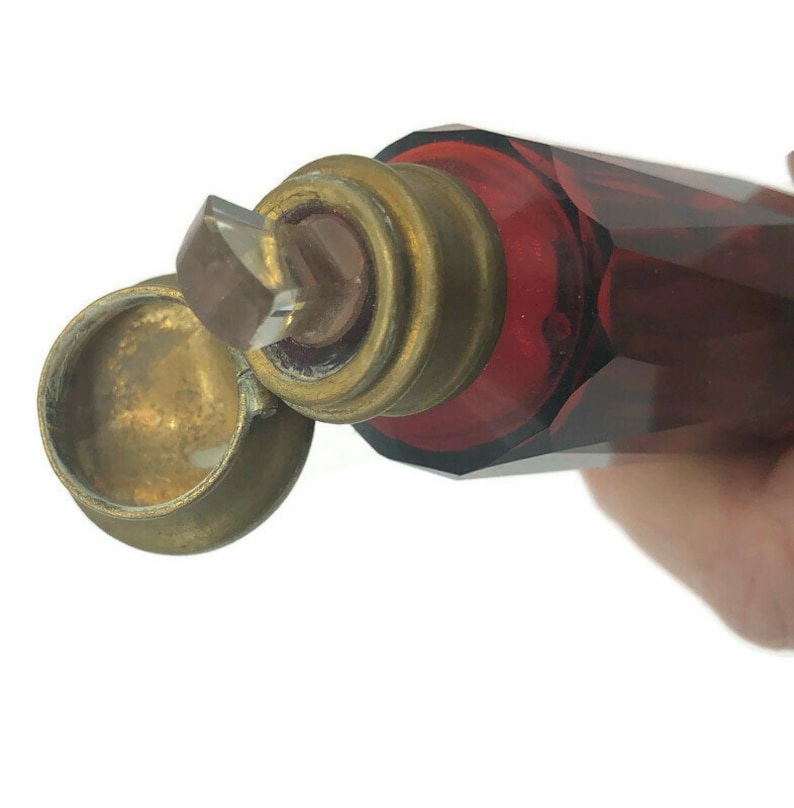 May include: A vintage glass bottle with a faceted design. The bottle is a deep red color and has a brass cap with a clear glass stopper. The bottle is open and the cap is off.