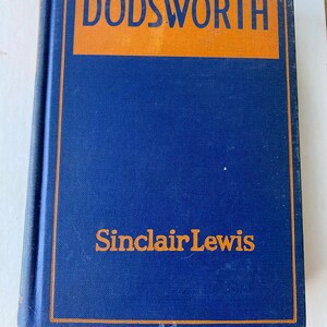 May include: A hardcover book titled "Dodsworth" by Sinclair Lewis. The book has a blue cover with orange lettering and a matching orange border. The book is closed and slightly worn.
