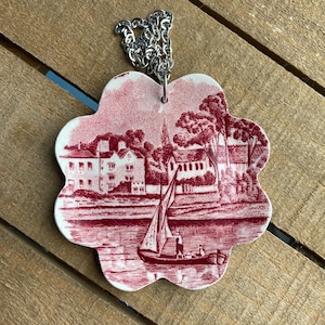 May include: A decorative pendant with a red and white toile design featuring a sailboat on a body of water, buildings, and trees. The pendant is flower-shaped and has a silver chain attached.