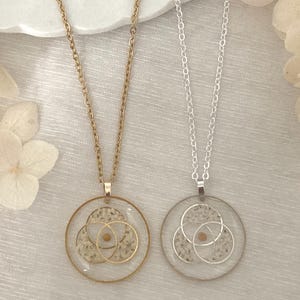 K&ouml;nnte beinhalten: Zwei Halsketten mit runden Anh&auml;ngern. Eine Halskette hat eine goldene Kette und einen Anh&auml;nger, die andere eine silberne Kette und einen Anh&auml;nger. Jeder Anh&auml;nger hat einen transparenten Harzkreis mit sich &uuml;berlappenden goldenen oder silbernen Kreisen und einem kleinen, runden, beigen Element.