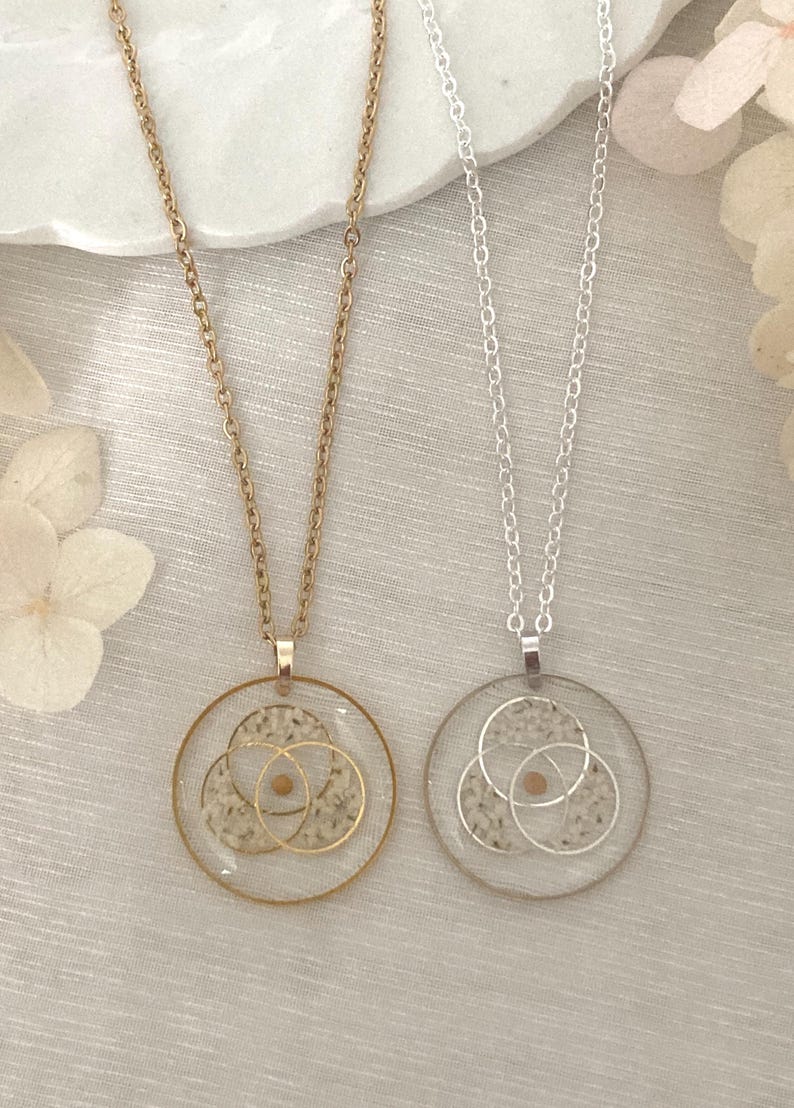 K&ouml;nnte beinhalten: Zwei Halsketten mit runden Anh&auml;ngern. Eine Halskette hat eine goldene Kette und einen Anh&auml;nger, die andere eine silberne Kette und einen Anh&auml;nger. Jeder Anh&auml;nger hat einen transparenten Harzkreis mit sich &uuml;berlappenden goldenen oder silbernen Kreisen und einem kleinen, runden, beigen Element.