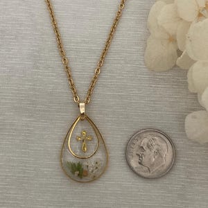 Pu&ograve; includere: Collana dorata con pendente a goccia contenente fiori secchi e una piccola croce dorata. Una moneta &egrave; posta accanto per riferimento alle dimensioni. La catena &egrave; a maglia delicata. Il pendente misura circa 2,5 cm di lunghezza.