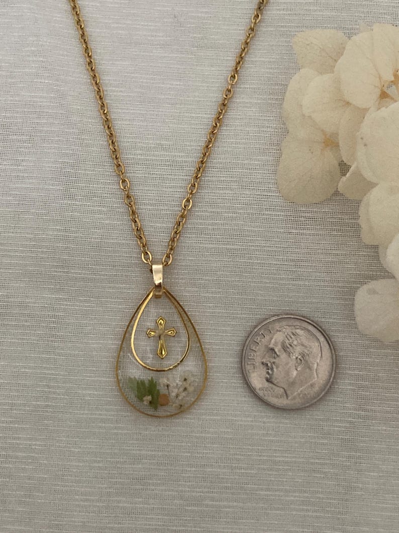 Pu&ograve; includere: Collana dorata con pendente a goccia contenente fiori secchi e una piccola croce dorata. Una moneta &egrave; posta accanto per riferimento alle dimensioni. La catena &egrave; a maglia delicata. Il pendente misura circa 2,5 cm di lunghezza.