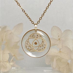 K&ouml;nnte beinhalten: Goldfarbene Halskette mit rundem Anh&auml;nger. Der Anh&auml;nger hat ein klares Harz, das ein Blumendesign mit einem goldfarbenen geometrischen Muster umschlie&szlig;t. Die Kette ist ebenfalls goldfarben, vor einem weichen wei&szlig;en Hintergrund.