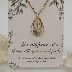 Pu&ograve; includere: Collana dorata con un ciondolo a goccia contenente una piccola croce e fiori secchi. La collana &egrave; appesa a una carta con la citazione "Come i fiori selvatici, fiorisce con grazia e fede."
