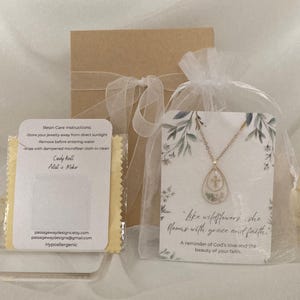 Pu&ograve; includere: Una collana color oro con un ciondolo a forma di goccia contenente fiori secchi, presentata con una confezione regalo e una carta con la scritta "like wildflowers, she blooms with grace and faith." Include anche un panno per la pulizia e le istruzioni per la cura.