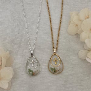 Pu&ograve; includere: Due collane con pendenti a goccia. Una &egrave; color argento, l'altra dorata. Ogni pendente presenta una piccola croce e fiori secchi. Le collane sono appese su uno sfondo di tessuto chiaro.