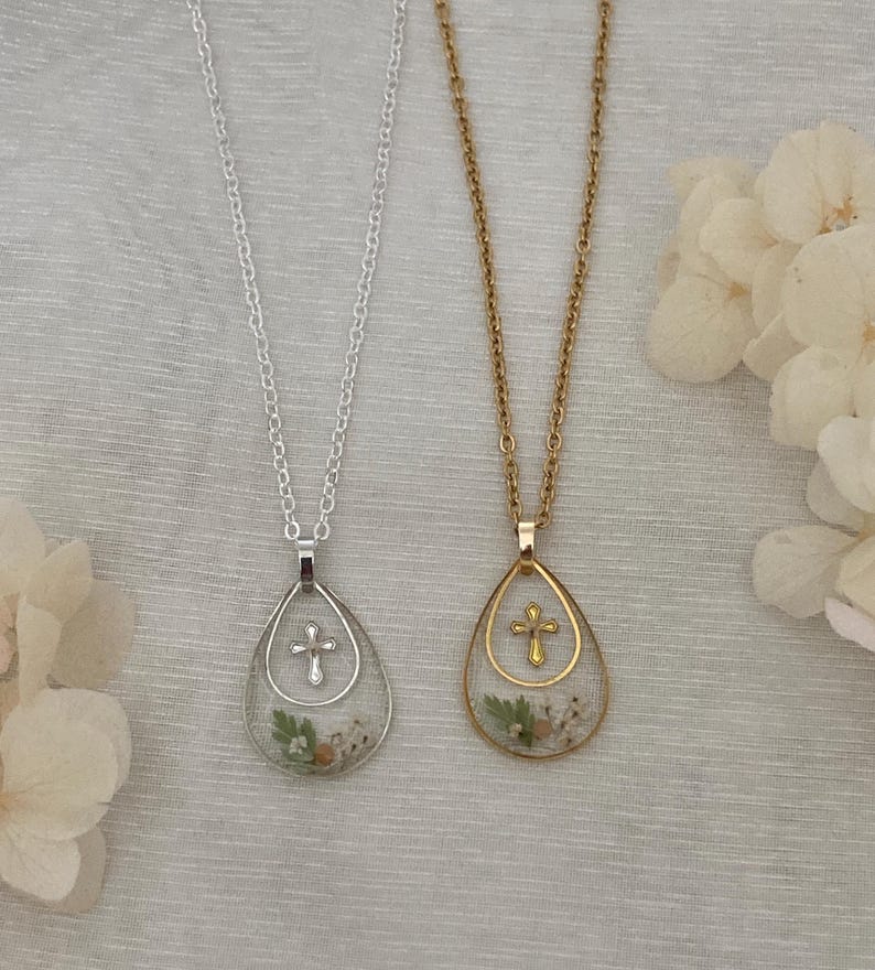 Pu&ograve; includere: Due collane con pendenti a goccia. Una &egrave; color argento, l'altra dorata. Ogni pendente presenta una piccola croce e fiori secchi. Le collane sono appese su uno sfondo di tessuto chiaro.
