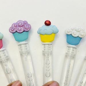 May include: Five bubble wands with cupcake toppers. Each wand has a clear plastic tube with bubbles inside. The cupcake toppers are in various colors, including pink, blue, yellow, and purple, with glitter and a red cherry on top.