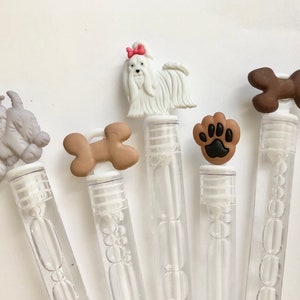 May include: Five clear plastic bubble wands with different dog-themed toppers. The toppers include a white dog with a pink bow, a brown bone, a gray dog, a brown bone, and a brown paw print.